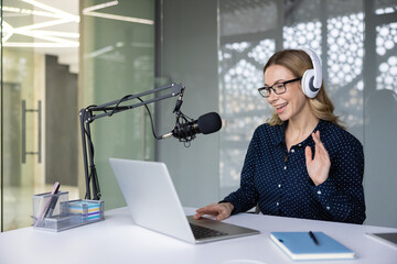 Woman wearing glasses and headphones, actively podcasting in an office setting, speaking into a microphone while using a laptop for live streaming or online communication
