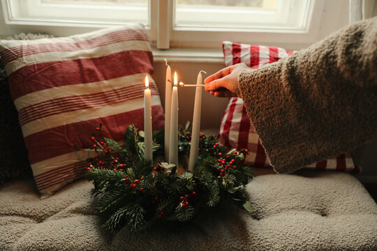 Female hand in beige wool sweater holding burning match lighting candle of advent wreath, red berries. Christmas still life. Winter interior, home decor. Blurred garden background. Cozy holiday mood.