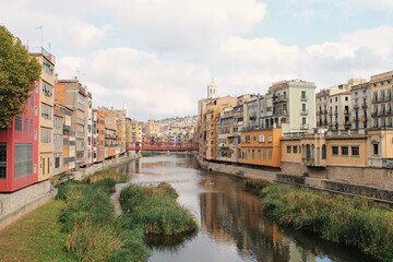 Elevated view of the Onyar River and historic multi-colored houses in Girona, Spain, featuring green reeds and a red metal bridge under a cloudy sky