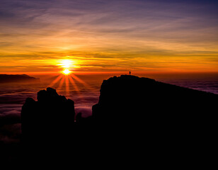 Silhouette of a solitary person on a mountain peak at sunrise above the clouds with brilliant orange and yellow sky and sunburst light rays