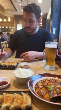 Caucasian man eating gyoza, mapo tofu, rice and beer with chopsticks in a Japanese restaurant