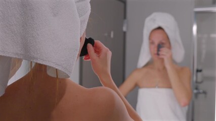 Woman with wet hair wrapped in towel massaging face using jade gua sha stone after shower to reduce puffiness and enhance natural glow