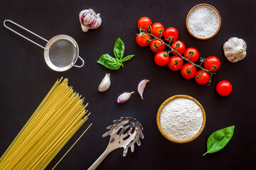 Flat lay of ingredients for cooking Italian pasta - uncooked spaghetti with tomatoes and basil leaves - on a black background, top view