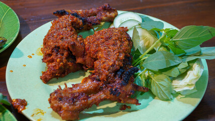 Two pieces of spicy grilled chicken thighs at one of the restaurants in Yogyakarta are served on a green plate and accompanied by basil leaves and cucumber as side dishes, placed on a brown table.