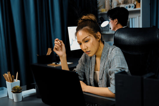 Serious face of thinking businesswoman focusing computer to report data analysis social media online project job with coworker working at back side at over late night time at modern office. Infobahn. - Powered by Adobe