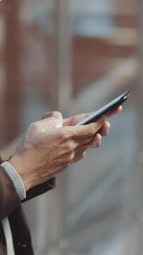 Vertical close-up shot of hands of anonymous young businessman texting colleague using smartphone with blurred background