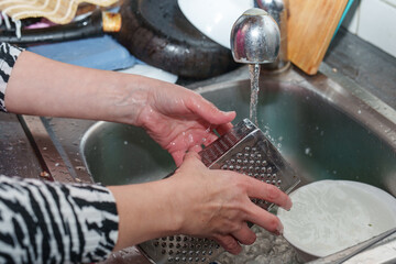 Hands washing a grater under running tap water inside a kitchen sink with other dishes after preparing a meal at dinner time