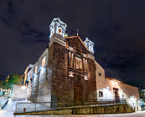 Stone facade of El Carmen Bajo church glows under night sky in Quito, Ecuador. Historic colonial building features wooden doors and twin bell towers in UNESCO World Heritage center