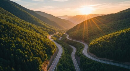 Winding mountain road at sunset, aerial view. Lush green forests surround a winding highway descending a valley. Sunlight radiates from a setting sun