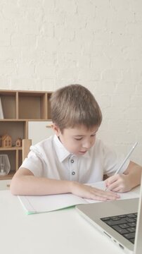 Schoolboy studies during online lesson on laptop, writing task in notebook at desk. Vertical video for education, e-learning and school themes