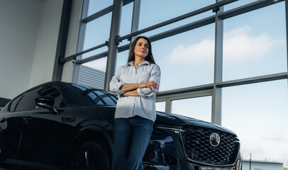 Big panoramic window is behind. Woman posing with a car in an auto showroom, modern dealership...