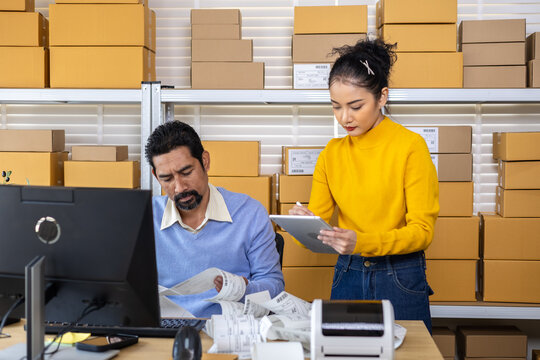 Small business couple analyzing printed labels, recording shipment updates by computer technology, coordinating inventory tasks, e-commerce operations with teamwork, and organizing warehouse planning