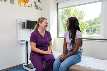 Medical professional in purple scrubs having a cheerful conversation with a young african american woman sitting on an examination table in a modern clinic