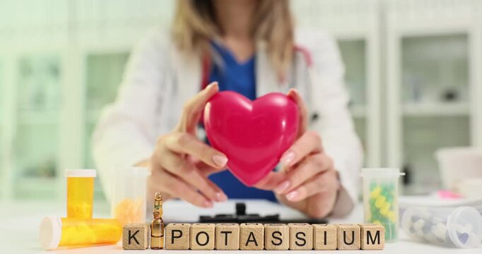 Wooden blocks spell word K-Potassium near red heart model. Cardiology woman holds heart shape explaining potassium role in rhythm to patient