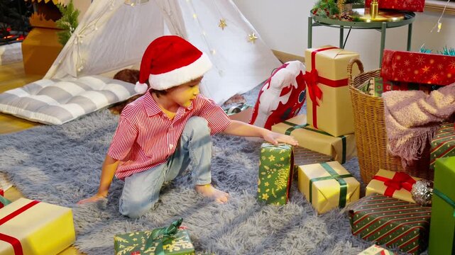 Asian Festive child santa hat opening wrapped gift rug beside teepee and holiday presents child santa hat reaches for wrapped christmas presents fluffy rug near cozy indoor tent creating warm festive