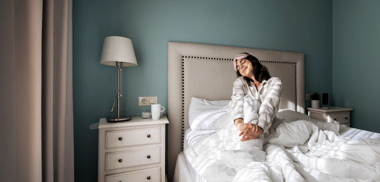 Woman in cozy pajamas stretches in a bright bedroom with soft bedding, a bedside lamp, and a serene atmosphere, embodying relaxation and morning comfort - Powered by Adobe