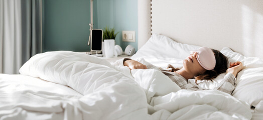 Woman wearing sleep mask is peacefully resting in a cozy bed with white bedding, surrounded by soft...