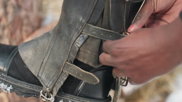 outdoor stable scene with tack inspection and repair, person adjusting leather strap on riding equipment outdoors in snow, carefully inspecting and fixing horse riding gear in snowy stable setting