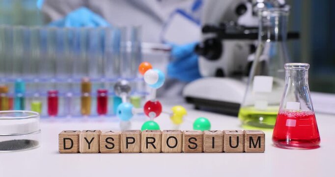 Wooden blocks spell word Dysprosium near molecular model and flasks. Laboratory worker studies solution checking dysprosium level under microscope