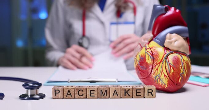Wooden blocks spell word Pacemaker beside anatomical heart model. Cardiology woman checks chart explaining pacemaker work for patient education