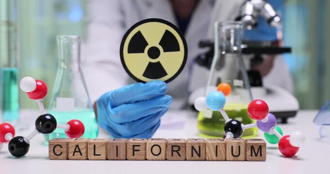 Wooden blocks spell word Californium near international radiation sign. Scientist person studies californium model recording results during experiment