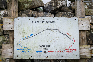 A weathered hand-painted sign marks the Pen-y-Ghent trail, showing routes to Horton Scar Lane and the Three Peaks path, mounted on rough wooden boards against a backdrop of stone and moss.