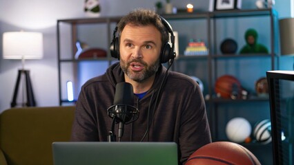 A man with headphones speaks into a microphone in a modern studio, surrounded by sports equipment and a computer, highlighting a dynamic podcasting environment.