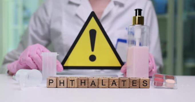 Woman laboratory technician sits at table holding warning sign in hands. Miniature wooden cubes on surface of desk clearly form word Phthalates