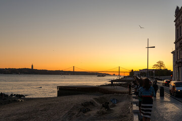Sunset over Tagus River and 25 de Abril Bridge in Lisbon, Portugal, with silhouettes of people walking along the waterfront promenade and Cristo Rei statue on distant hill.