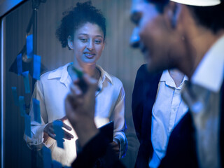 Diverse group of business professionals collaborating on transparent glass board. Writing with sticky notes for strategic project planning and technology development in the office late at night.