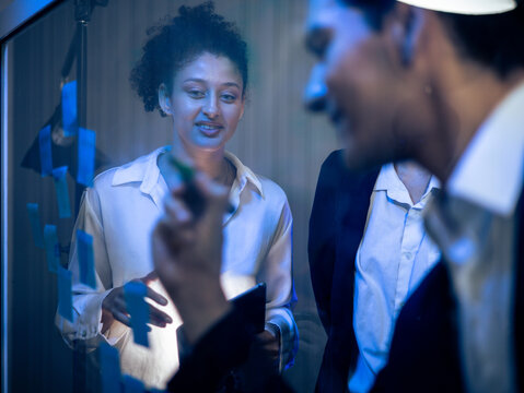 Diverse group of business professionals collaborating on transparent glass board. Writing with sticky notes for strategic project planning and technology development in the office late at night.