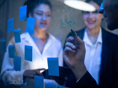 Team of business professionals is focused on strategic development in tech office. Writing on glass board with sticky notes for innovative technology planning and successful project execution.