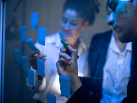 Diverse group of business professionals collaborating on transparent glass board. Writing with sticky notes for strategic project planning and technology development in the office late at night.