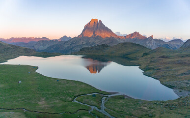 Aerial view of a mountain lake reflecting a peak at sunrise with green meadows and distant mountains