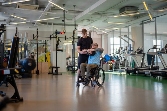 Personal sports trainer assists disabled sportsman in wheelchair with medicine ball workout to build upper body strength. Athlete undergoing rehabilitation in sports center with professional coach.