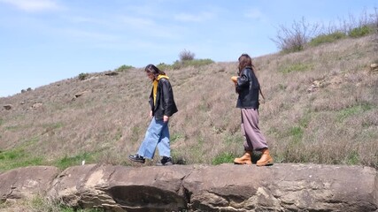 Cheerful girl friends walk leisurely through quiet countryside. Smiling woman in casual attire uses modern smartphone to photograph cloud-streaked sky