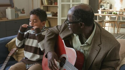 Talented Black boy child playing harmonica with enjoyment while granddad accompanying on acoustic guitar sitting on couch in living room