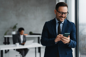 A businessman in a modern office standing near a window and using his smartphone and in the background, an African businesswoman is seated at a desk working on a laptop