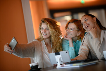 Three young businesswomen take a selfie at a table in an office space during their lunch break