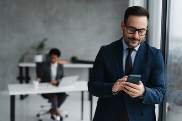 A businessman in a modern office standing near a window and using his smartphone and in the background, an African businesswoman is seated at a desk working on a laptop
