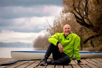 Sporty active senior man sitting on the jetty by the river next to his kayak