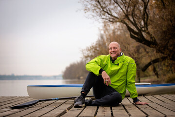 Sporty active senior man sitting on the jetty by the river next to his kayak