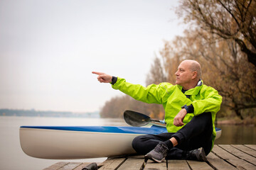 Sporty active senior man sitting on the jetty by the river next to his kayak