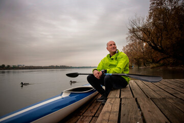 Sporty active senior man sitting on the jetty by the river next to his kayak