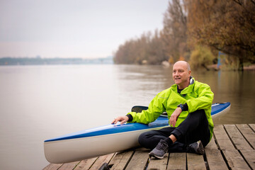 Sporty active senior man sitting on the jetty by the river next to his kayak