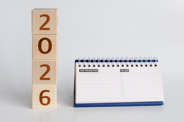 Wooden blocks with "2026" written on them stand vertically next to spiral notepad with to-do list and priority section for effective planning and organization.