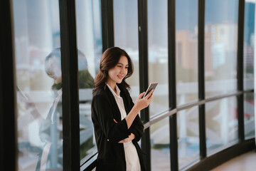 Fototapeta premium A professional woman standing by large windows, smiling while working on a laptop in a bright modern office environment.
