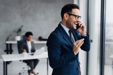 A businessman in a modern office standing near a window and using his smartphone and in the background, an African businesswoman is seated at a desk working on a laptop
