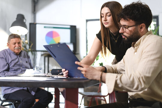 Business discussion takes place in a modern office setting with a man reviewing a document and a woman providing assistance during a meeting with another colleague