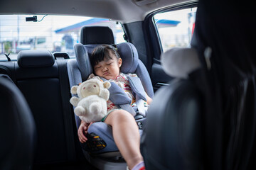 A cute little Asian girl is sleeping in the backseat of a car and hugging a teddy bear,Little girl with teddy bear, tablet and headphones in the car during travel on vacation.